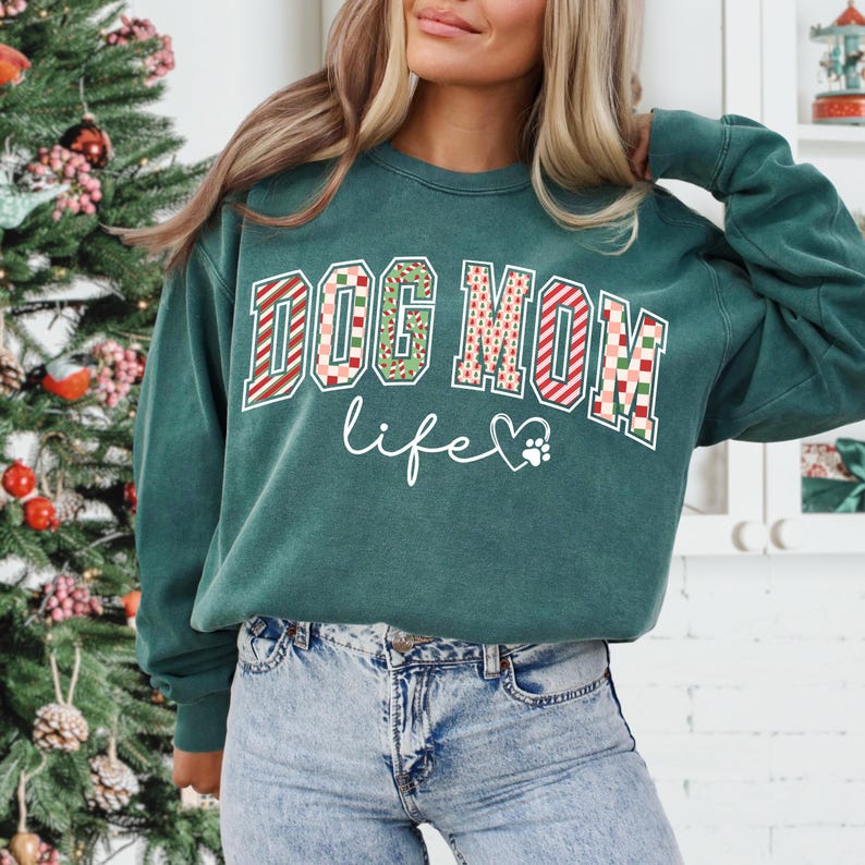 Może przedstawiać: Zielona bluza z napisem "DOG MOM life". Słowa "DOG MOM" są w odważnym, wzorzystym designie z czerwonymi i zielonymi akcentami. "life" jest napisane delikatnym skryptem z sercem w kształcie łapy.