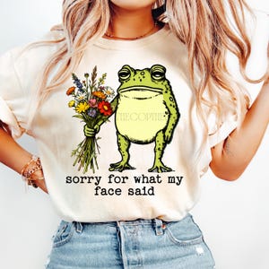 May include: Beige t-shirt featuring a green frog holding a bouquet of colorful flowers. The frog has a grumpy expression. The text "sorry for what my face said" is printed below the frog.