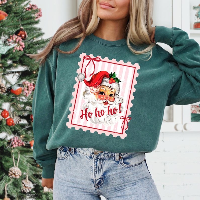 May include: A teal green sweatshirt featuring a vintage-style Santa Claus graphic within a pink and white striped frame. The Santa illustration includes a red hat, white beard, and the text "Ho ho ho!" in red. The sweatshirt is a casual style.
