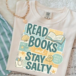 以下が含まれることがあります： クリーム色のTシャツ。テキスト「READ BOOKS and STAY SALTY」は、ティールと黄色で書かれています。デザインには、貝殻、開いた本、ヒトデのイラストが含まれています。ペンダント付きのゴールドネックレスが見えます。