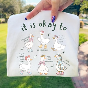 Pode incluir: Camiseta branca com gansos de desenho animado e frases encorajadoras como "it is okay to have hard days" e "be yourself." Os gansos usam acess&oacute;rios e s&atilde;o cercados por estrelas. A camiseta &eacute; segurada por uma pessoa.