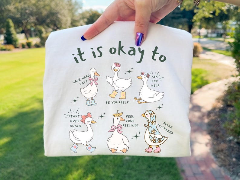 Pode incluir: Camiseta branca com gansos de desenho animado e frases encorajadoras como "it is okay to have hard days" e "be yourself." Os gansos usam acess&oacute;rios e s&atilde;o cercados por estrelas. A camiseta &eacute; segurada por uma pessoa.