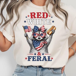 K&ouml;nnte beinhalten: Cremefarbenes T-Shirt mit einem Waschb&auml;ren, der eine herzf&ouml;rmige Sonnenbrille, eine Schleife und ein Halstuch in Rot, Wei&szlig; und Blau tr&auml;gt. Der Waschb&auml;r h&auml;lt eine amerikanische Flagge und einen Hotdog. Der Text auf dem Shirt lautet "RED WHITE & FERAL."