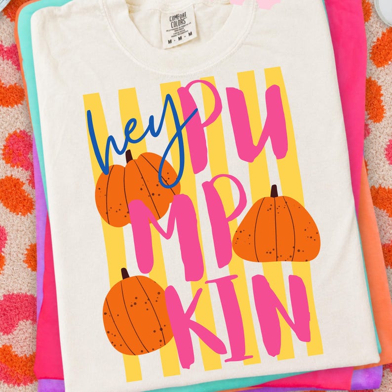 May include: Cream-colored t-shirt with the text "hey PUMPKIN" in blue and pink, featuring orange pumpkin illustrations and yellow striped background. The shirt is folded on top of other colorful shirts.