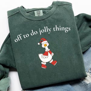 Könnte beinhalten: Ein dunkelgrünes Sweatshirt mit Rundhalsausschnitt und dem weißen Schriftzug "off to do jolly things" über einer Grafik einer weißen Gans, die eine rote Weihnachtsmütze, einen roten Umhang mit weißem Saum und rote Stiefel trägt. Mit Comfort Colors Etikett.