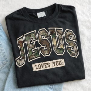 K&ouml;nnte beinhalten: Schwarzes T-Shirt mit dem Wort "JESUS" in gro&szlig;en Buchstaben im Camouflage-Muster mit wei&szlig;er Umrandung. Darunter steht der Satz "LOVES YOU" in einem rechteckigen, abgenutzten Feld.