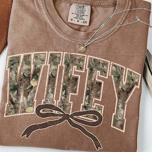 May include: A brown t-shirt with the word "WIFEY" in a camouflage pattern across the front. A brown bow is below the word. A silver necklace with a pendant is on the shirt. The shirt has a Comfort Colors tag.
