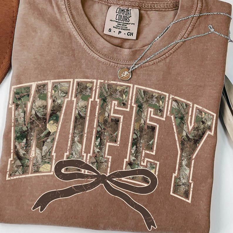 May include: A brown t-shirt with the word "WIFEY" in a camouflage pattern across the front. A brown bow is below the word. A silver necklace with a pendant is on the shirt. The shirt has a Comfort Colors tag.