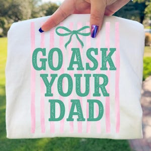 Puede incluir: Camiseta blanca con rayas verticales rosas y el texto "GO ASK YOUR DAD" en verde azulado. Un lazo verde azulado est&aacute; en la parte superior. La camiseta se sostiene sobre un fondo verde.