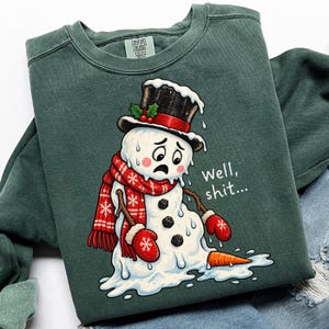 Könnte beinhalten: Ein dunkelgrüner Rundhals-Pullover mit einer Grafik eines schmelzenden Schneemanns, der einen schwarzen Zylinder, einen roten Schal mit weißen Schneeflocken und rote Fäustlinge trägt. Der Schneemann schaut traurig und daneben steht Well, shit...