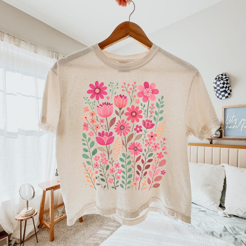 May include: Cream-colored t-shirt featuring a vibrant floral design. The graphic showcases a variety of pink flowers, green leaves, and other botanical elements. The shirt is hanging on a wooden hanger.