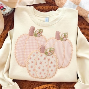 May include: A cream-coloured crewneck sweatshirt featuring three appliquéd pumpkins on the front. The pumpkins are crafted from fabric with pink gingham, pink striped, and pink floral patterns, each finished with decorative stitching and lace trim.