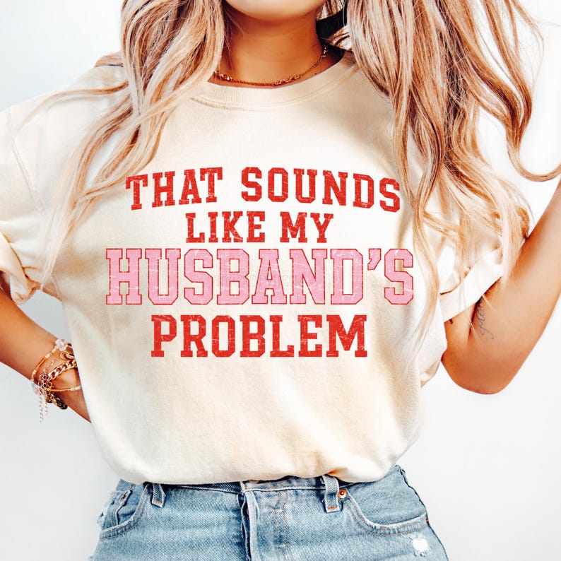 May include: Cream-colored t-shirt with the text "THAT SOUNDS LIKE MY HUSBAND'S PROBLEM" in red and pink block letters. The shirt is a casual style, suitable for everyday wear.