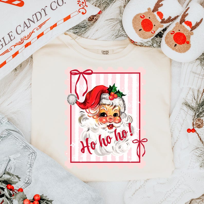 May include: Cream-colored sweatshirt featuring a vintage Santa Claus graphic within a red frame. Santa wears a red hat and says "Ho ho ho!" in a festive font. The design includes pink and white stripes and decorative bows, evoking a holiday theme.