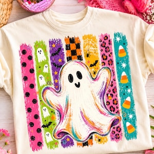 May include: A cream-colored crewneck t-shirt featuring a large central white ghost graphic. The design includes vertical brushstroke stripes in pink, green, purple, orange, and teal, decorated with ghosts, bats, spiders, webs, leopard print, and candy corn.