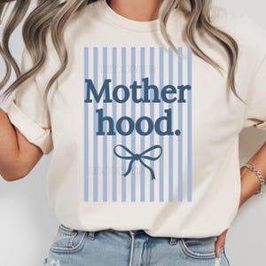 K&ouml;nnte beinhalten: Cremefarbenes T-Shirt mit dem Text "Motherhood" in Marineblau, vor einem hellblau gestreiften Hintergrund. Unter dem Text befindet sich eine marineblaue Schleife. Das Shirt hat kurze &Auml;rmel und einen Rundhalsausschnitt.