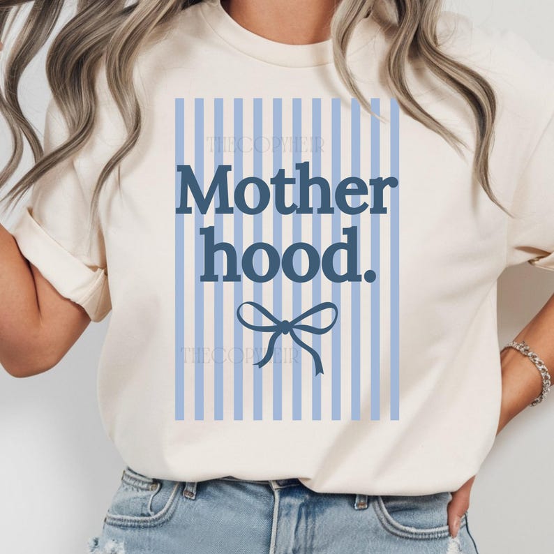 K&ouml;nnte beinhalten: Cremefarbenes T-Shirt mit dem Text "Motherhood" in Marineblau, vor einem hellblau gestreiften Hintergrund. Unter dem Text befindet sich eine marineblaue Schleife. Das Shirt hat kurze &Auml;rmel und einen Rundhalsausschnitt.