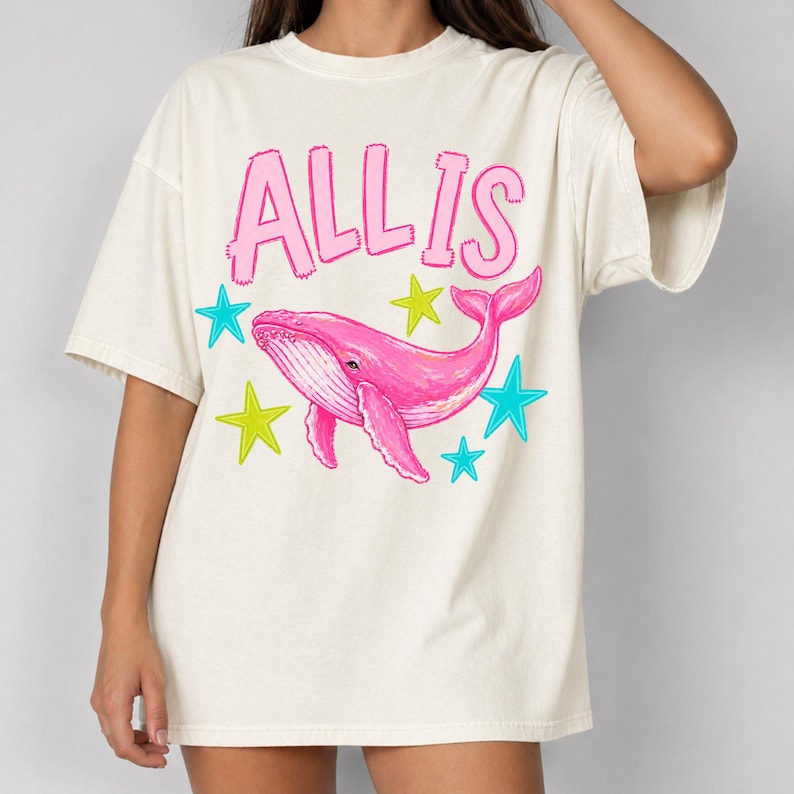 May include: Cream-colored t-shirt featuring the text "ALL IS" in pink above a pink whale illustration. The whale is surrounded by teal and yellow stars. The shirt has short sleeves and a relaxed fit.