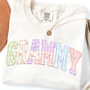 May include: Cream-colored t-shirt with the word "GRAMMY" in colorful patchwork letters. The letters feature floral and gingham patterns. A gold necklace with a pendant rests on the shirt. The shirt has a Comfort Colors tag.