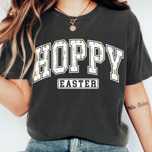 May include: Dark gray t-shirt with the word "HOPPY" in a white outlined, collegiate-style font, filled with a pattern of pastel-colored Easter bunnies. Below, the word "EASTER" is in a white, rectangular box. The shirt is a comfortable, casual style.