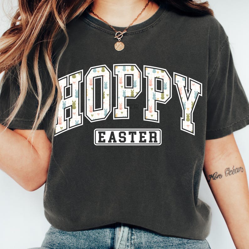 May include: Dark gray t-shirt with the word "HOPPY" in a white outlined, collegiate-style font, filled with a pattern of pastel-colored Easter bunnies. Below, the word "EASTER" is in a white, rectangular box. The shirt is a comfortable, casual style.