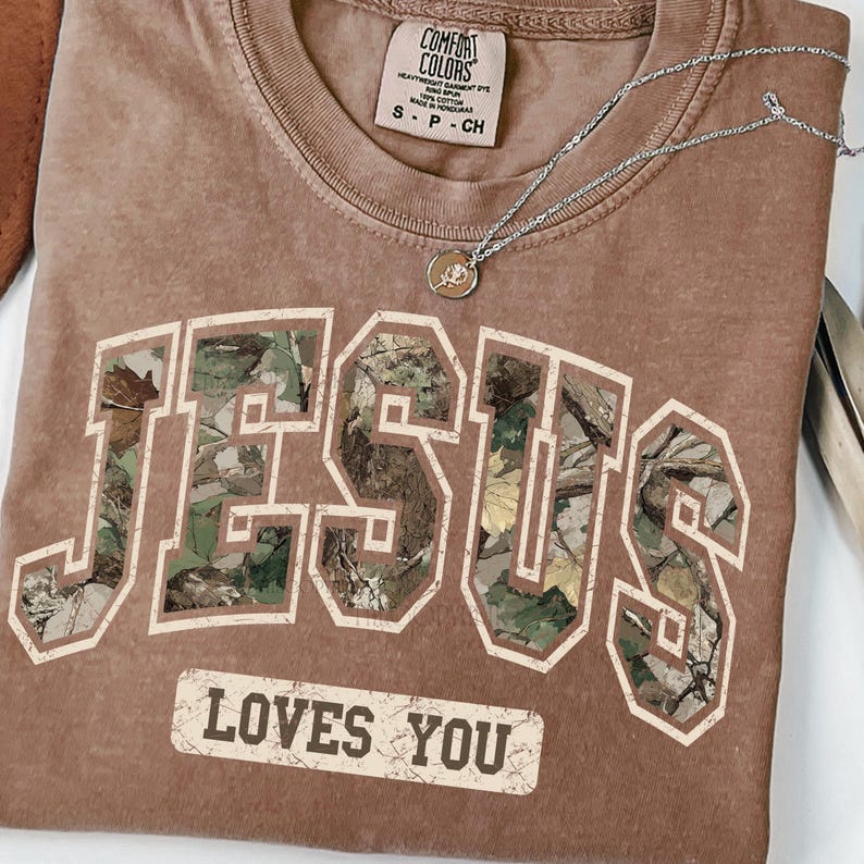 K&ouml;nnte beinhalten: Ein braunes T-Shirt mit dem Wort "JESUS" in gro&szlig;en, mit Tarnmuster gef&uuml;llten Buchstaben. Darunter ist der Text "LOVES YOU" auf einem rechteckigen Etikett aufgedruckt. Eine silberne Halskette mit Anh&auml;nger liegt auf dem Shirt.