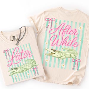 May include: Two cream-colored t-shirts with a graphic of an alligator wearing sunglasses. One shirt says "Later" and the other says "After While" in pink script. Both shirts have a striped background in teal and pink accents.
