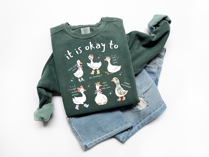 Pode incluir: Camisola verde escuro com o texto "it is okay to" e ilustra&ccedil;&otilde;es de gansos. Os gansos usam acess&oacute;rios e a camisola &eacute; combinada com cal&ccedil;&otilde;es de ganga azuis.