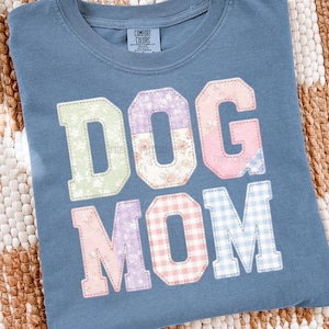 Può includere: Una maglietta blu Comfort Colors con la scritta "DOG MOM" in un design patchwork. Le lettere sono in vari modelli, tra cui floreale, gingham e colori pieni. La maglietta è realizzata in un materiale morbido.