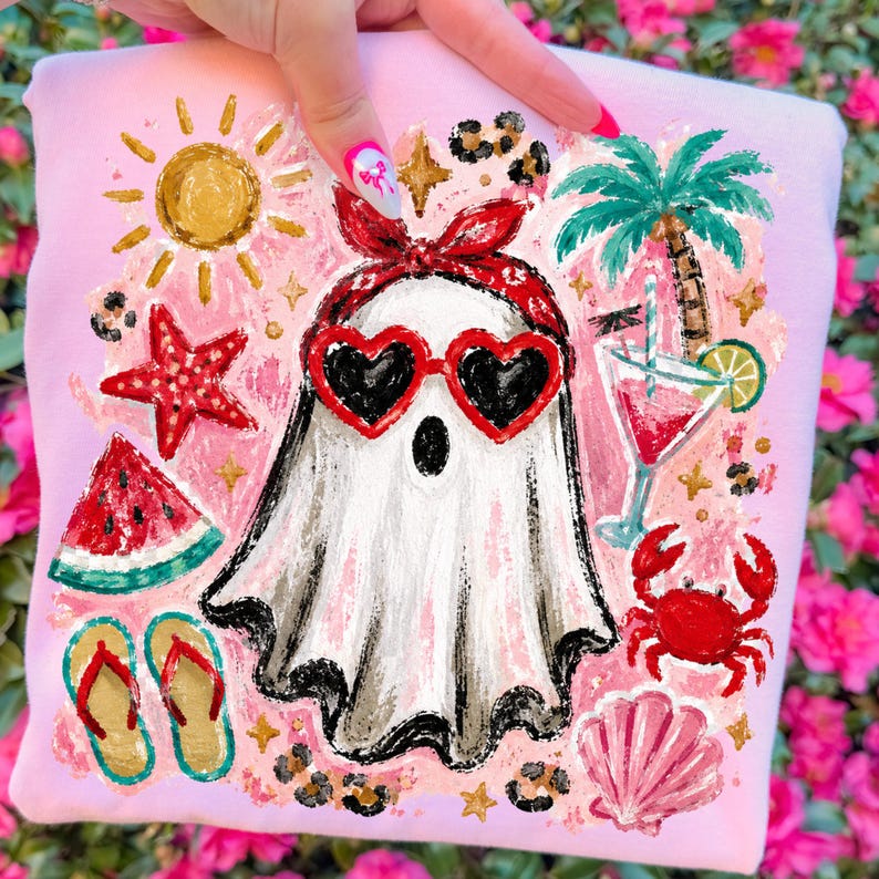 May include: Pink sweatshirt with a whimsical beach-themed design. A white ghost wearing heart-shaped sunglasses and a red bandana is surrounded by a sun, starfish, watermelon, flip-flops, palm tree, cocktail, crab, and seashell.