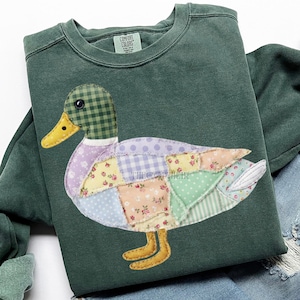 May include: Green sweatshirt featuring a patchwork duck design. The duck has a checkered head, a yellow beak, and a body composed of various patterned fabric patches in pastel colors. The sweatshirt has a crew neck and long sleeves.