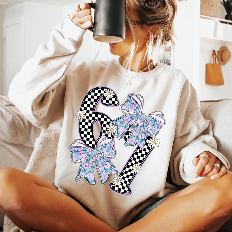 May include: Cream-colored sweatshirt with a large graphic featuring the numbers "67" in a black and white checkered pattern. The numbers are adorned with floral bows in shades of blue, pink, and green, and accented with small white daisies.