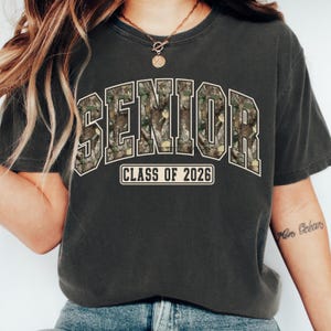 Pu&ograve; includere: T-shirt grigio scuro con la scritta "SENIOR" in un motivo mimetico, bordata di bianco. Sotto, il testo "CLASS OF 2026" in un riquadro bianco. La maglietta &egrave; in stile casual.