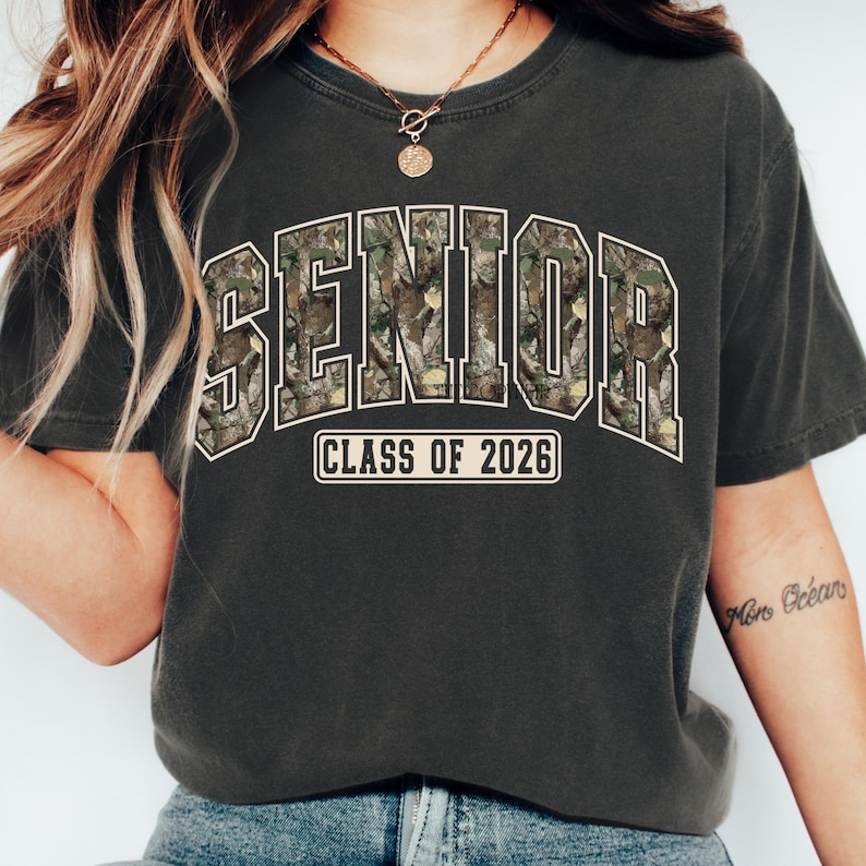 Pu&ograve; includere: T-shirt grigio scuro con la scritta "SENIOR" in un motivo mimetico, bordata di bianco. Sotto, il testo "CLASS OF 2026" in un riquadro bianco. La maglietta &egrave; in stile casual.