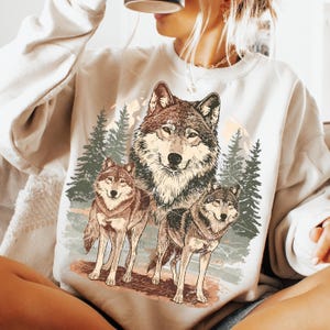 Puede incluir: Una sudadera crema con un gr&aacute;fico detallado de tres lobos en un entorno forestal. El dise&ntilde;o incluye pinos y una cordillera en el fondo, con una est&eacute;tica vintage y desgastada.