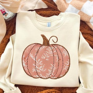 Peut inclure: Sweat-shirt crème avec un motif de citrouille brodée. La citrouille est rose clair avec un motif floral blanc et une tige marron. Le motif est souligné d'une couture marron.