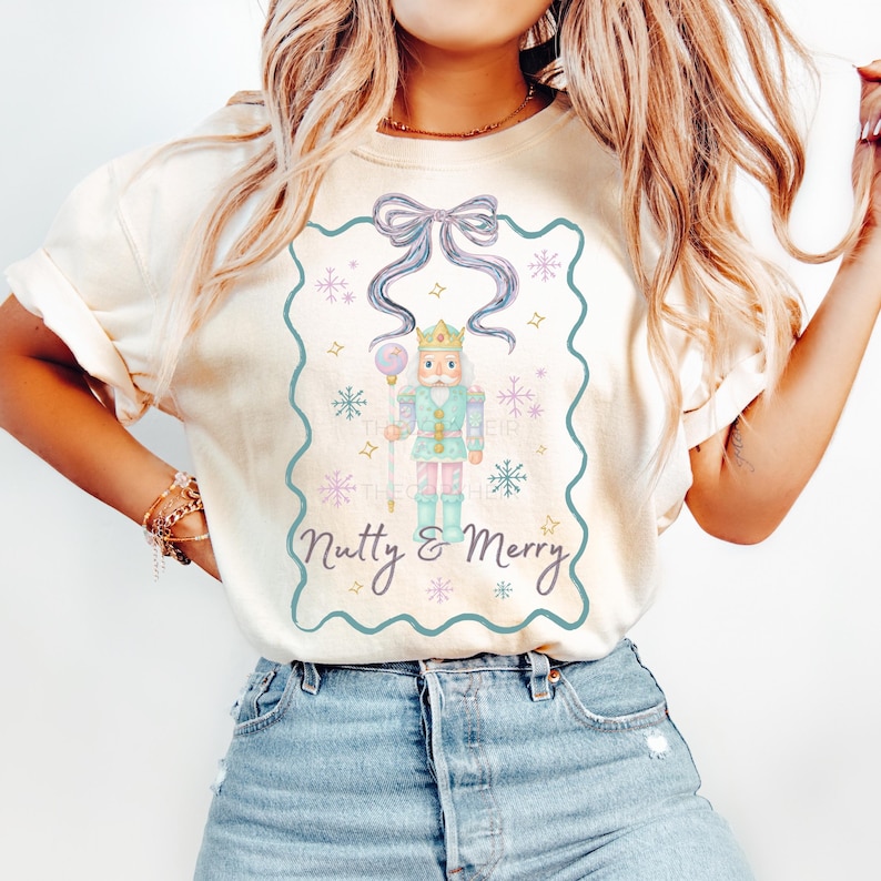 K&ouml;nnte beinhalten: Cremefarbenes T-Shirt mit einer Aquarellillustration eines Nussknackersoldaten in einem t&uuml;rkisfarbenen Rahmen. Das Design umfasst eine blaue Schleife, Schneeflocken und den Text "Nutty & Merry". Ein l&auml;ssiges, festliches Kleidungsst&uuml;ck.