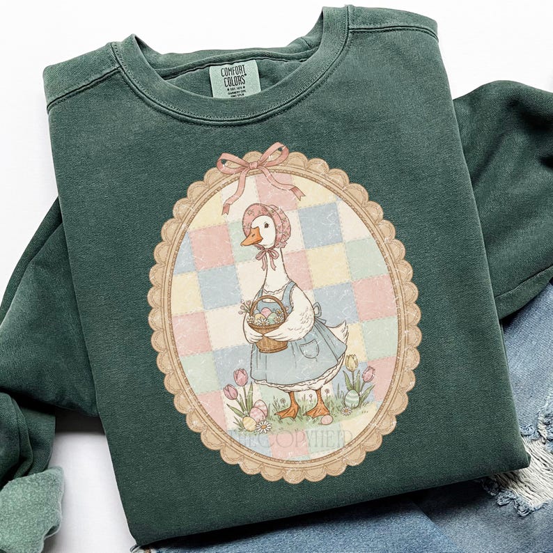 Puede incluir: Una sudadera Comfort Colors verde oscuro con una ilustraci&oacute;n enmarcada de un ganso con gorro y delantal, sosteniendo una cesta de huevos de Pascua. La ilustraci&oacute;n est&aacute; ambientada sobre un fondo de patchwork pastel, con tulipanes y huevos a los pies del ganso.