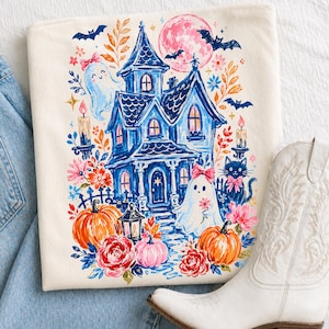 May include: A cream-colored t-shirt featuring a colorful graphic print of a blue haunted house, two ghosts with pink bows, a black cat, orange pumpkins, and various flowers. The design includes a pink moon, bats, and lit candles in a painterly style.