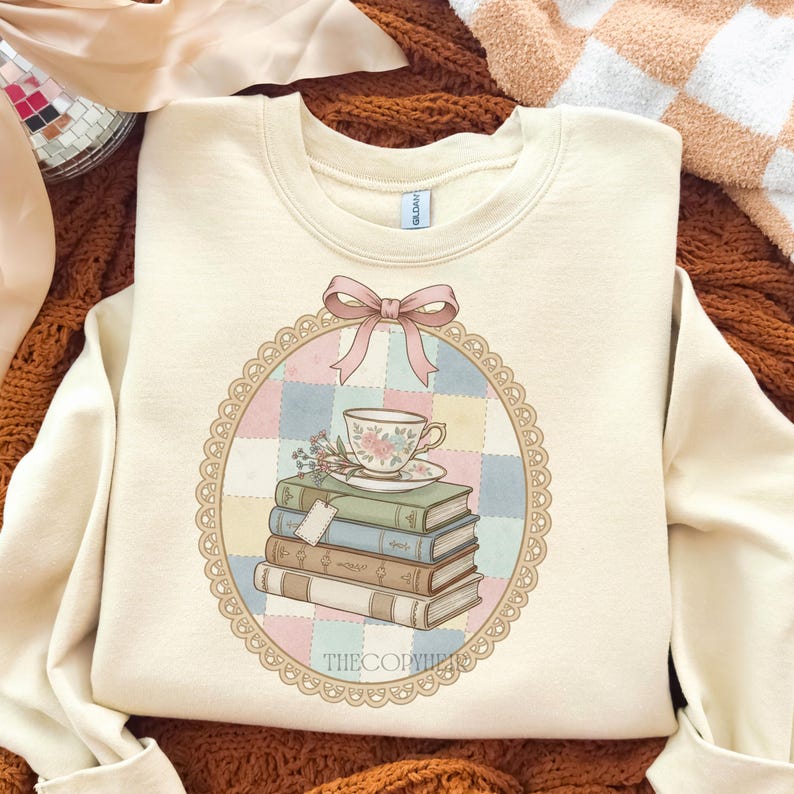 May include: Cream-coloured sweatshirt with a round graphic featuring a stack of books, a teacup, and a pink bow. The graphic is surrounded by a lace-like border and a pastel patchwork background. The text "THECOPYWRITER" is at the bottom.
