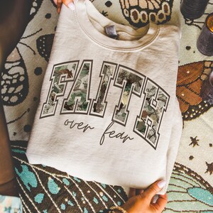 May include: A light beige sweatshirt with the word "FAITH" in large, block letters filled with a camouflage pattern. Below the word "FAITH" is the phrase "over fear" in a cursive font. The sweatshirt is folded.