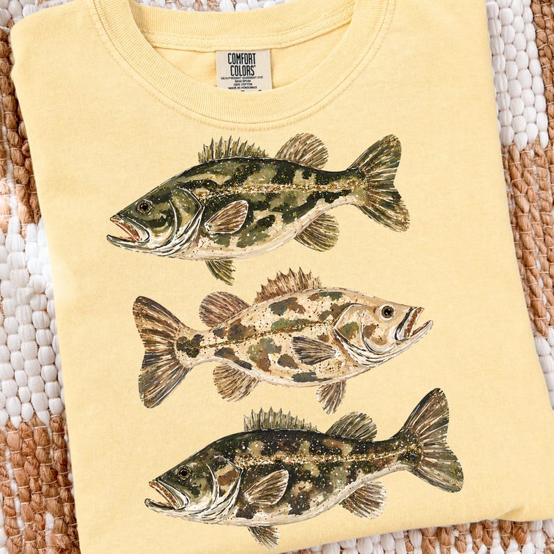 May include: Yellow t-shirt featuring three detailed illustrations of fish. The fish have camouflage-like patterns in shades of green, brown, and tan. The shirt has a crew neck and a tag that reads "Comfort Colors."