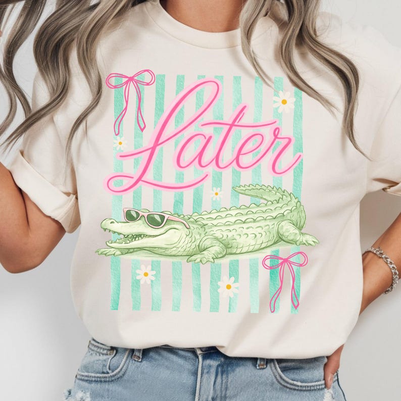 K&ouml;nnte beinhalten: Cremefarbenes T-Shirt mit einer Grafik, die ein gr&uuml;nes Alligator mit Sonnenbrille, das Wort "Later" in rosa Schrift und einen Hintergrund aus mintgr&uuml;nen Streifen und G&auml;nsebl&uuml;mchen zeigt. Rosa Schleifen akzentuieren das Design.