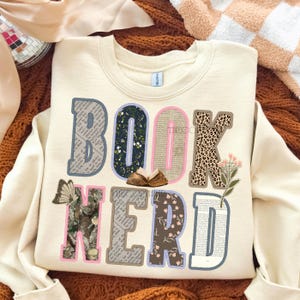 May include: Cream-colored sweatshirt with the words "BOOK NERD" in a colorful, layered design. The letters feature patterns like floral, leopard print, and book pages. A butterfly and small open book are also included.