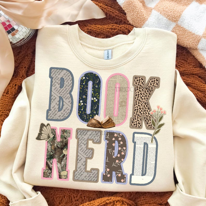 May include: Cream-colored sweatshirt with the words "BOOK NERD" in a colorful, layered design. The letters feature patterns like floral, leopard print, and book pages. A butterfly and small open book are also included.