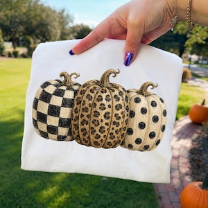 May include: White fabric featuring three decorative pumpkins. One pumpkin has a black and white checkered pattern, the center pumpkin has a leopard print, and the third pumpkin has black polka dots. The pumpkins have brown stems.