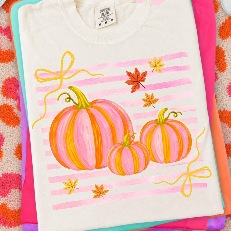 May include: Cream-colored t-shirt featuring a watercolor design of three pink and orange pumpkins with yellow stems and leaves. Pink horizontal stripes and yellow ribbons accent the pumpkins. The shirt is folded on top of other colorful shirts.