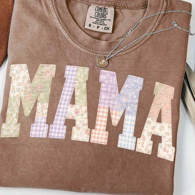 May include: Brown t-shirt with the word "MAMA" in patchwork letters. The letters feature floral and gingham patterns in pastel colors. A silver necklace with a pendant rests on the shirt.