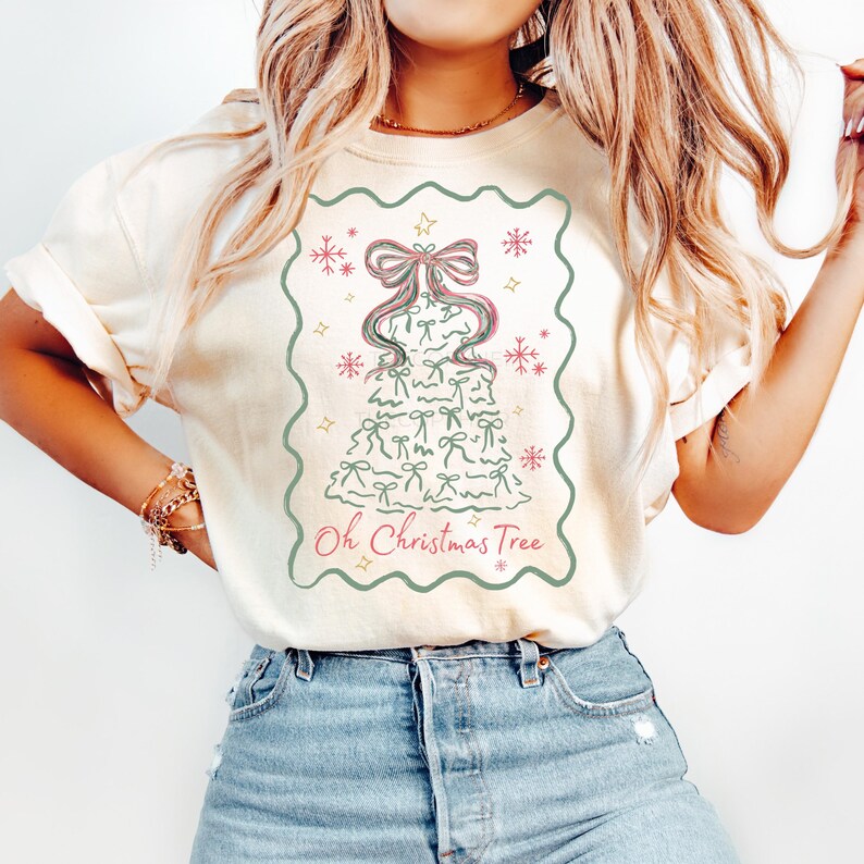 Peut inclure: T-shirt cr&egrave;me avec un motif de sapin de No&euml;l orn&eacute; de n&oelig;uds et de flocons de neige. Le motif est encadr&eacute; d'une bordure verte et porte l'inscription "Oh Christmas Tree". Le t-shirt est port&eacute; par une personne.
