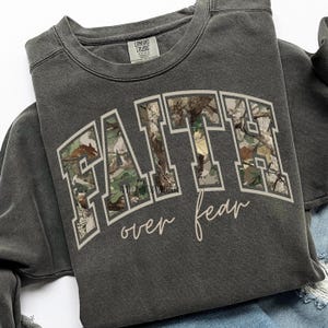 May include: Dark gray sweatshirt with the words "FAITH" in a block letter design filled with a camouflage pattern. The words "over fear" are written in a cursive font below the word "FAITH".
